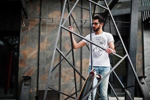 gata stil arabisk man i glasögon med longboard poserade inuti metall pyramid konstruktion. foto