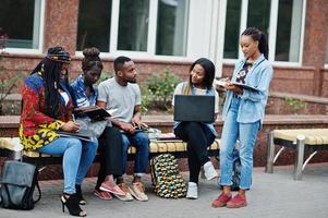 grupp på fem afrikanska högskolestudenter som spenderar tid tillsammans på campus på universitetsgården. svarta afro vänner studerar vid bänken med skolsaker, bärbara datorer anteckningsböcker. foto