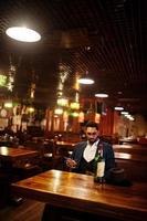 stilig välklädd arabisk man med ett glas whisky och cigarrhåll mobiltelefon, poserade på puben. foto