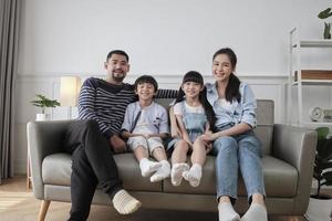 porträtt av asiatisk thailändsk familj, vuxen pappa, mamma med små barn lycka hem bor tillsammans, tittar på kameran, fritid på soffan i vita rummet, härlig helg och välbefinnande inhemsk livsstil. foto