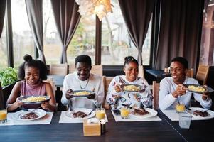 glada afrikanska vänner sitter, chattar på café och äter mat. grupp av svarta folk som träffas i restaurangen och äter middag. de håller tallrikar med pommes frites. foto