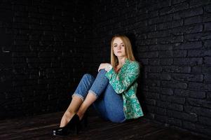 snygg blond tjej i jacka och jeans mot tegelsvart vägg på studio. foto