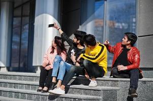 grupp asiatiska vänner som sitter på trappor utomhus mot modern byggnad och gör selfie via telefon. foto