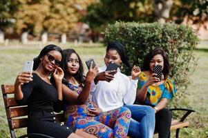grupp av fyra afroamerikanska flickor som sitter på bänken utomhus med mobiltelefoner vid händerna och gör selfie. foto