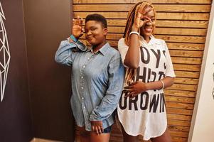 två afrikanska kvinnor i snygga vardagskläder poserar mot trävägg och visar ok fingrar. foto