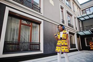 snygga afroamerikanska kvinnor i gul jacka poserade och hörlurar på gatan mot modern byggnad med varm dryck i engångspappersmugg. foto