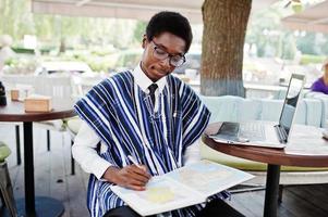 afrikansk man i traditionella kläder och glasögon sitter bakom laptop på utomhuscafé och tittar på kartan över Afrika och Ghana på sin anteckningsbok. foto