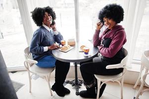 två lockigt hår afrikansk amerikansk kvinna bär tröjor sitter vid bordet café, äter croissant och dricker te. foto