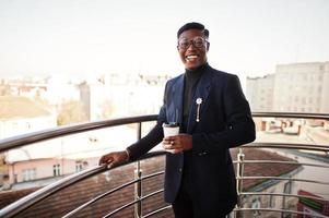 fashionabla afrikansk amerikansk man i kostym och glasögon med en kopp kaffe vid händerna poserade på balkongen på kontoret. foto