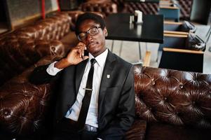 affärsafrikansk amerikansk man bär på svart kostym och glasögon sitter på kontoret och talar i telefon. foto