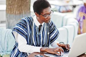 afrikansk man i traditionella kläder och glasögon sitter bakom laptop på utomhuscafé och arbetar. foto