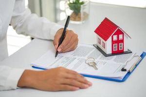 en kvinna skriver på ett kontrakt om att köpa ett hus med en fastighetsmäklare. modellhus med nycklar på bordet. foto