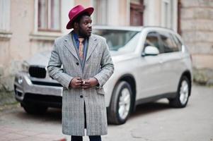 snygg afrikansk amerikansk man modell i grå kappa, jacka slips och röd hatt mot silver business suv bil. foto
