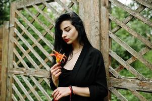 sensuell tjej helt i svarta, röda läppar. goth dramatisk kvinna håller orange lilja blomma. foto