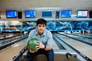 snygg asiatisk man i jeansskjorta stående vid bowlinghall med boll till hands. foto