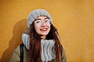porträtt av brunett flicka i grå halsduk och hatt, glasögon vid kallt väder med solsken mot orange vägg av gamla hus. gratis kopieringsutrymme. foto
