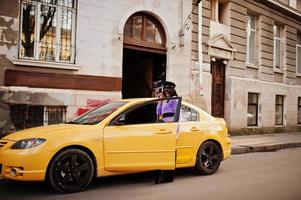 afrikansk amerikansk kvinna på violett klänning och mössa poserade på gul bil. foto