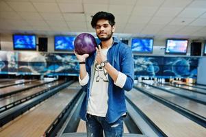 elegant skägg asiatisk man i jeansskjorta stående vid bowlinghall med boll till hands. foto