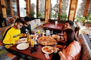 grupp asiatiska vänner som äter pizza under fest på pizzeria. glada indianer har roligt tillsammans, äter italiensk mat och sitter på soffan. foto