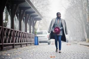 snygg afrikansk amerikansk man modell i grå kappa, jacka slips och röd hatt poserade på dimmigt väder gata. foto