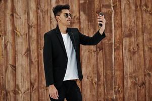 Casual ung indisk man i svart kavaj och solglasögon poserade mot träbakgrund och gör selfie på mobiltelefonen. foto