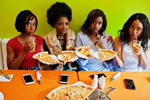 fyra unga afrikanska flickor i ljusa färgade restaurang med pizza skivor på tallrik och juice. foto