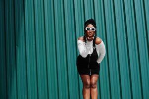 afrikansk kvinna i jeansoverallkjol, vita solglasögon mot grön stålvägg, skickar luftkyss. foto