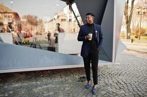 otroligt snygg afrikansk amerikansk man bär på blå kavaj med brosch, svart polotröja och glasögon poserade på gatan. fashionabla svart kille med kopp kaffe. foto