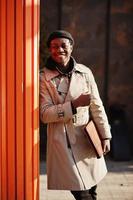stilig afrikansk amerikansk man poserar utanför i svart hatt och beige kappa med mapp i handen. foto