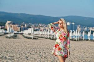 blond kvinna som bär klänning står på sandstranden vid havet och njuter av utsikten över solnedgången. foto