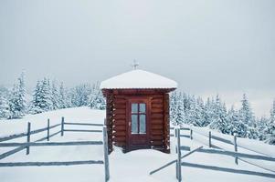 trä litet kyrkkapell vid tallar täckta av snö på berget chomiak. vackra vinterlandskap av Karpaterna, Ukraina. frost natur. foto