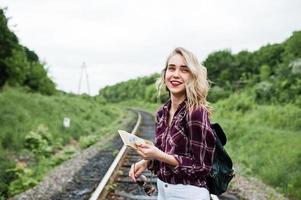 porträtt av en vacker blond tjej i tartanskjorta som går på järnvägen med kartan i händerna. foto