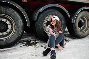 brunett snygg casual tjej i mössa och solglasögon sitter mot lastbil hjul. foto