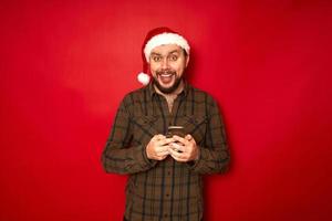 en glad upprymd man i en jultomteshatt med en telefon i händerna gläds åt mottagen sensation. isolerad på röd studio bakgrund. koncept - nytt år, applikation, internet, kommunikation foto