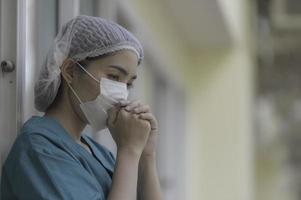 trött deprimerad kvinnlig asiatisk skrubbsköterska bär ansiktsmask blå uniform sitter på sjukhusgolvet, ung kvinna läkare stressad av hårt arbete foto