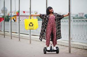 vacker afrikansk amerikansk kvinna som använder segway eller hoverboard. svart flicka med gul duk eco väskor återvinning symbol. foto