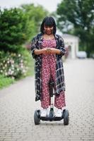 vacker afrikansk amerikansk kvinna som använder segway eller hoverboard. svart flicka på dubbla hjul självbalanserande elektrisk skoter med mobiltelefon till hands. foto