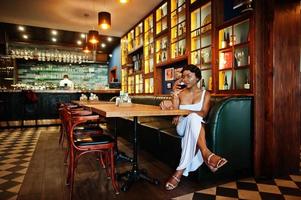 afrikansk amerikansk kvinna, retro frisyr i vit klänning på restaurang med ett glas vin. foto