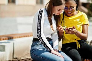 vit kaukasisk flicka och svart afroamerikan tittar tillsammans på mobiltelefonen. världsenhet, raskärlek, förståelse i tolerans och samarbete mellan raser och mångfald. foto
