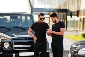 två asiatiska bröder man bär på helt svart poserade nära suv bilar och tittar på mobiltelefon. foto