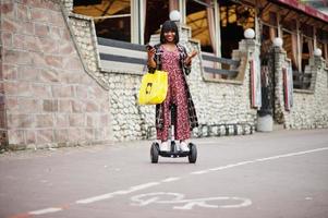 vacker afrikansk amerikansk kvinna som använder segway eller hoverboard. svart flicka med gul duk eco väskor återvinning symbol. foto