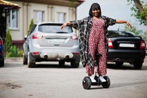 vacker afrikansk amerikansk kvinna som använder segway eller hoverboard. svart flicka på dubbla hjul självbalanserande elektrisk skoter mot bilparkering. foto