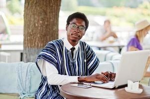 afrikansk man i traditionella kläder och glasögon sitter bakom laptop på utomhuscafé och arbetar. foto