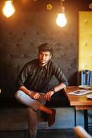 självsäker ung indisk man i svart skjorta sitter på café. foto