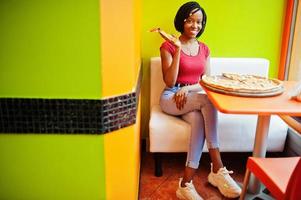 afrikansk kvinna med pizza sitter på ljusa färgade restaurang. foto