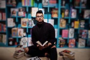 lång smart arab student man, bära på violett turtleneck och glasögon, på biblioteket sitter mot bokhyllor. foto