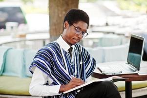 afrikansk man i traditionella kläder och glasögon sitter bakom laptop på utomhuscafé och skriver något på sin anteckningsbok. foto