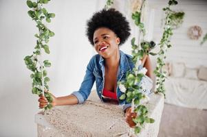 attraktiv afrikansk amerikansk kvinna med afro hår slitage på kjol och jeans jacka, poserade på vita rummet på swing. moderiktig svart modell. foto