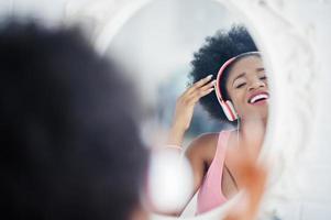 ung afroamerikansk kvinna tittar i spegeln och lyssna på musik i hörlurar. foto