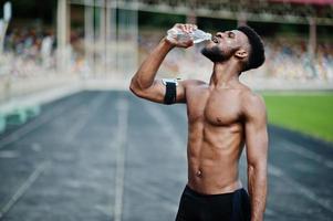 afrikansk amerikansk manlig idrottare sexig sport bar överkropp man med löpande sportarmfodral för mobiltelefon, poserade på stadion och dricksvatten från flaska. foto